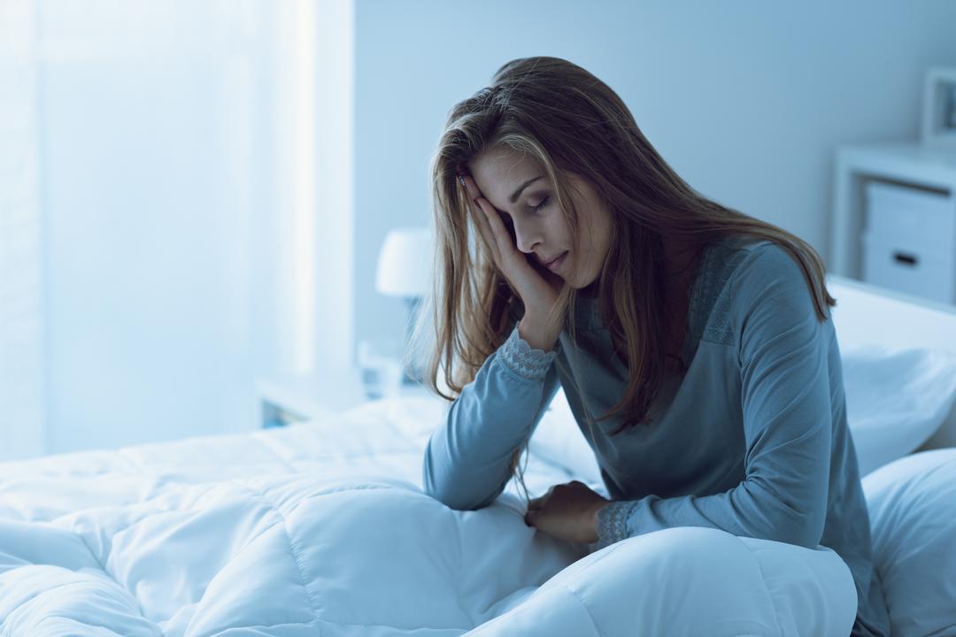 Eine erschöpfte Frau sitzt auf ihrem Bett und stützt ihren Kopf mit der Hand, was Müdigkeit und Schlafprobleme symbolisiert.