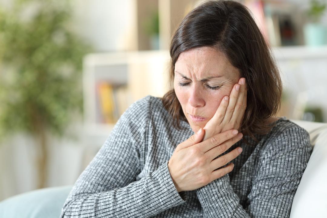 Eine Frau hält sich schmerzverzerrt die Wange, um Zahnschmerzen zu lindern. Dieses Bild illustriert die Qual von Zahnschmerzen und die Dringlichkeit, schnelle Hilfe zu finden.